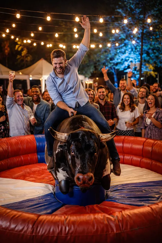 Mekanisk tjur som aktivitet på fest och event – kvällsstämning med ljusslingor och publik som hejar på deltagare i uppblåsbar arena.